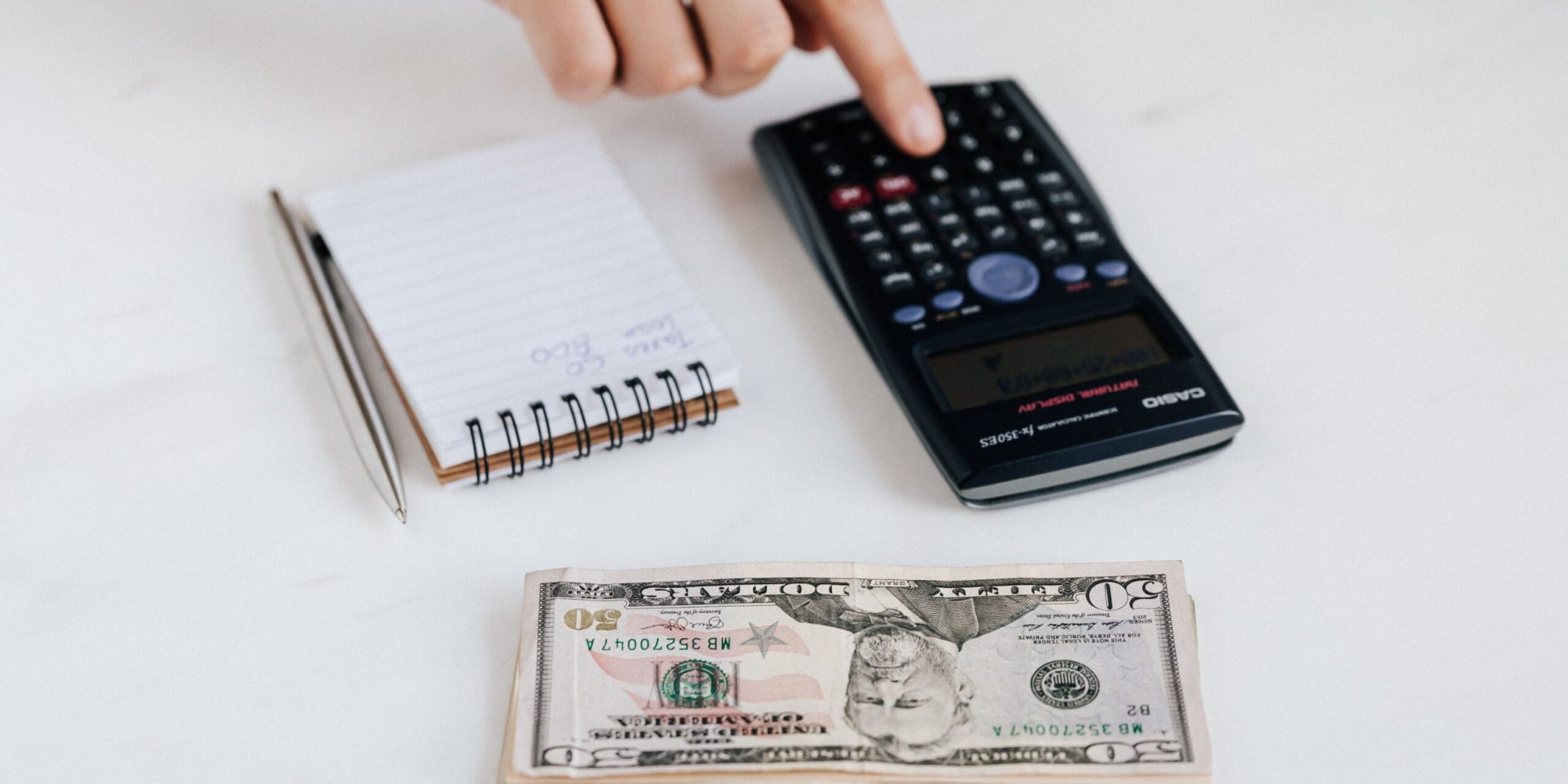 Top view of crop faceless economist touching calculator button while counting income near heap of paper bills and small notebook with pen