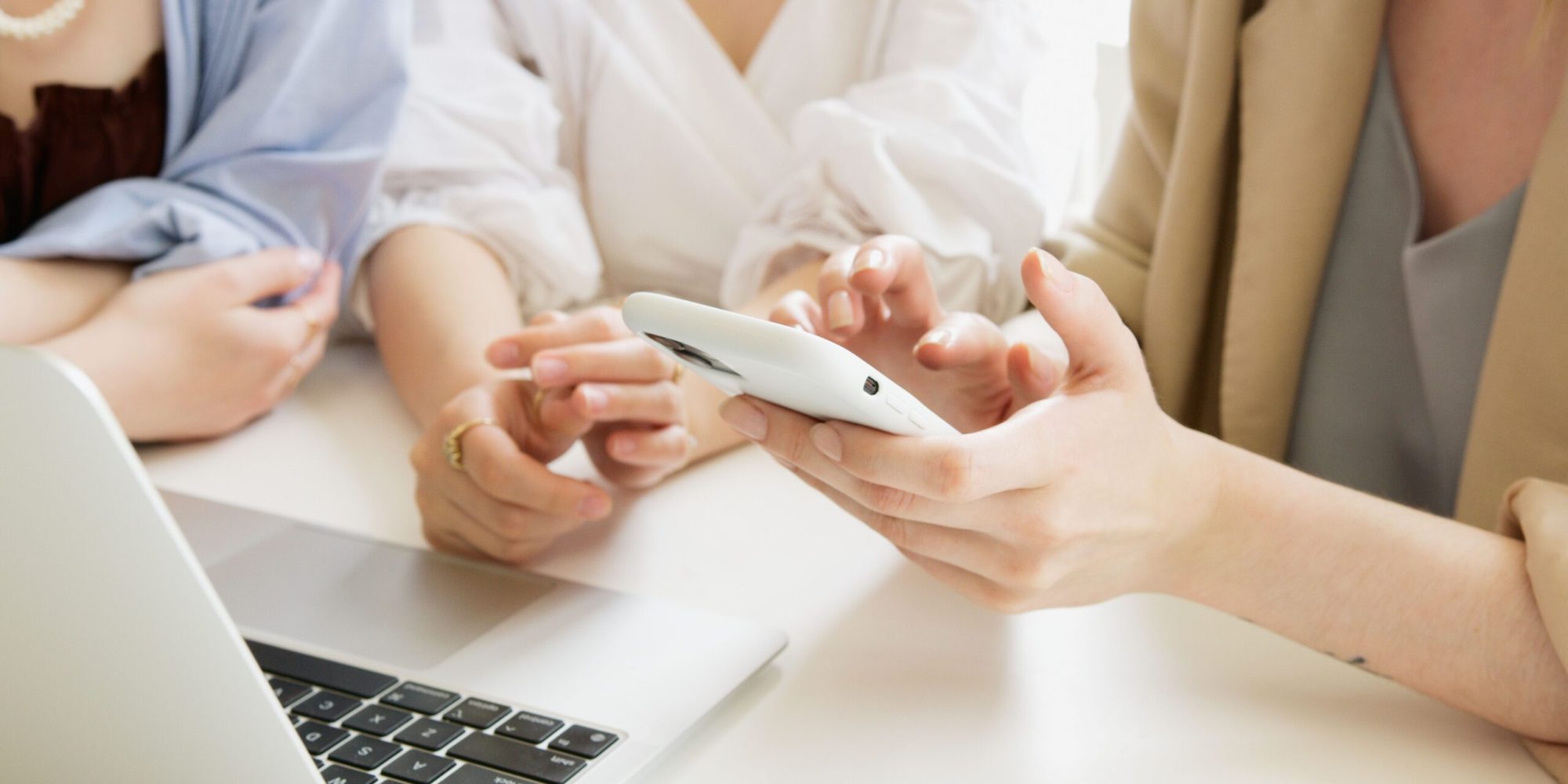 Three women collaborate using a smartphone and laptop in a professional setting.